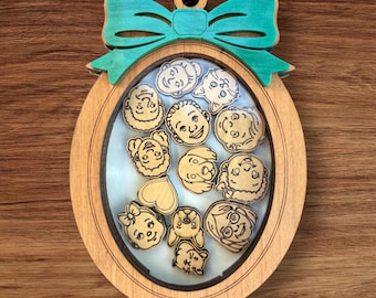 Pode incluir: Enfeite oval de madeira com um laço verde-azulado e rostos de desenhos animados no interior. O enfeite de madeira clara tem um pequeno orifício para pendurar. Os rostos são gravados na madeira.