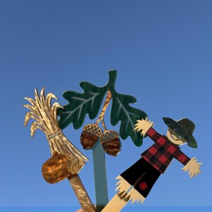 May include: A collection of wooden fall-themed decorations. Includes a bundle of wheat with a pumpkin, a green oak leaf with acorns, and a scarecrow in a red and black plaid shirt. The background is a clear blue sky.