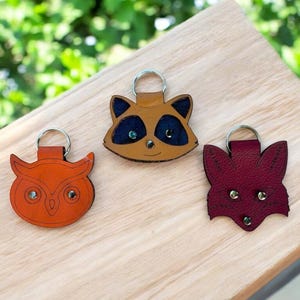 May include: Three leather animal keychains: an orange owl, a raccoon with blue accents, and a burgundy fox. Each keychain has a silver ring. The keychains are on a light wood surface.