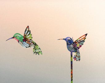 May include: Two decorative hummingbirds. One is green with red and yellow details, the other is blue with orange and yellow accents. One hummingbird is attached to a decorative stick.