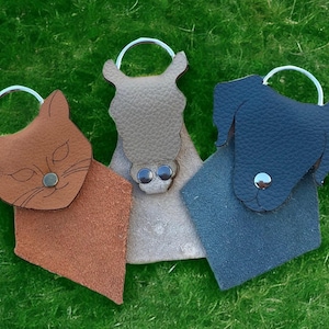 May include: Three animal-shaped leather keychains with metal rings. One is a brown cat, one is a beige horse, and one is a dark blue dog. Each keychain features a snap button closure and a textured surface, designed to hold keys securely.