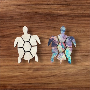 May include: Two turtle-shaped wooden puzzles. One turtle is plain light-colored wood with visible grain, while the other features a vibrant, multi-colored iridescent pattern with swirls of blue, purple, pink, and green across its segmented shell.
