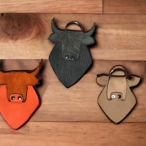 Puede incluir: Tres llaveros de cuero con forma de toro en colores naranja, verde oscuro y beige. Cada llavero presenta una superficie texturizada, una anilla metálica en la parte superior y dos remaches de metal plateado como orificios nasales.
