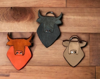 Pode incluir: Três porta-chaves de couro em forma de touro nas cores laranja, verde-escuro e bege. Cada porta-chaves apresenta uma superfície texturizada, um anel metálico no topo e dois rebites de metal prateado como narinas no focinho.