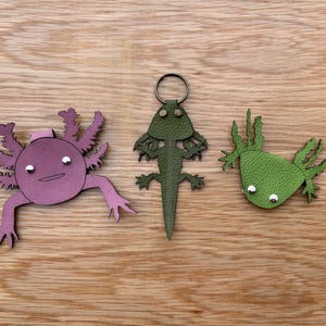 May include: Three leather keychains shaped like axolotls. One is pink, one is green, and one is green with a silver ring.