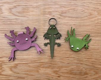 Pode incluir: Três chaveiros de couro em forma de axolotl. Um é rosa, outro é verde e outro é verde com um anel prateado.