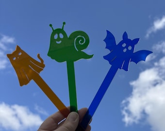 Pode incluir: Três estacas de jardim de acrílico coloridas: um gato laranja, um caracol verde e um morcego azul. Cada estaca tem um design caprichoso e é segurada contra um céu azul brilhante com nuvens brancas fofas. Estas estacas decorativas adicionam um toque lúdico a qualquer jardim.