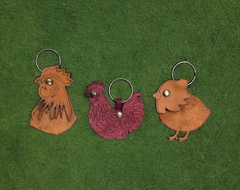 Pode incluir: Três porta-chaves de couro em forma de aves. À esquerda, uma cabeça de galo castanha, ao centro, uma galinha magenta com penas gravadas, e à direita, um pintainho castanho. Cada um possui um anel de metal prateado e um pequeno rebite.