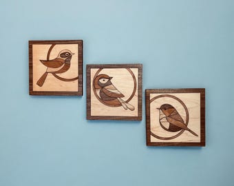 May include: Three square wooden wall art pieces featuring inlaid bird designs. Each panel displays a different bird silhouette crafted from various wood tones, set against a light wood background and framed by a dark wood border.