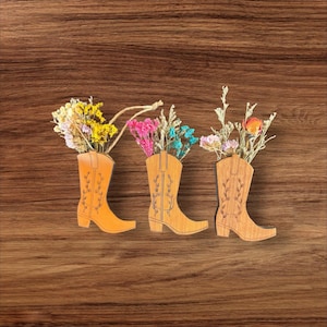 Puede incluir: Tres adornos de pared de madera con forma de bota de vaquero, cada uno con un patrón de enredadera floral grabado con láser. Las botas están rellenas con pequeños ramos de flores secas en tonos amarillo, rosa, azul y naranja, con un cordel.