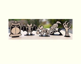 Pode incluir: Um conjunto de cinco recortes de madeira em preto e bege apresentando um cérebro num frasco, um esqueleto de morcego, um esqueleto de rato segurando um globo ocular, um esqueleto de pássaro num ramo e uma mão esquelética sobre bases.