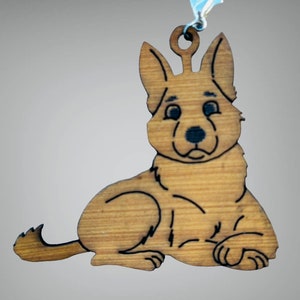 Op de afbeelding: Houten ornament in de vorm van een Duitse herdershond. De hond ligt met opstaande oren. Het ornament is gemaakt van lichtbruin hout met zwarte details en een kleine lus om op te hangen.