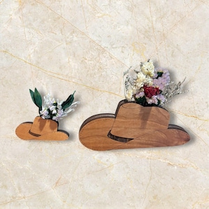 Puede incluir: Dos floreros de pared de madera con forma de sombrero de vaquero en diferentes tamaños. Cada sombrero presenta un acabado de veta de madera natural y contiene un pequeño arreglo de flores secas, incluyendo pétalos blancos, rosas y rojo oscuro.