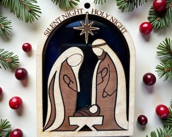 Pode incluir: Um ornamento de Natal de madeira em forma de arco com uma cena de natividade cortada a laser, com figuras em tons de castanho sobre um fundo azul escuro. O texto SILENT NIGHT HOLY NIGHT está gravado no topo, com uma estrela acima.