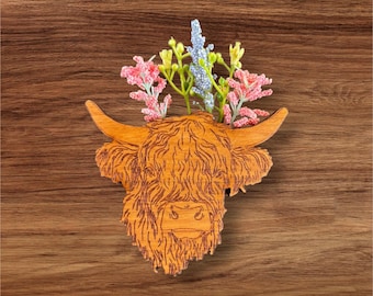 Pode incluir: Um recorte de madeira de uma cabeça de vaca das terras altas com um arranjo floral no topo. As flores são rosa, azuis e verdes.