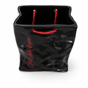May include: A black ceramic vase shaped like a shopping bag with a glossy finish and crinkled texture. The front features the red text Saks Fifth Avenue. It includes two red rope handles threaded through silver-toned metal grommets at the top.