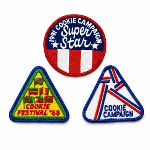 May include: Three embroidered patches: a circular red, white, and blue patch reading 1991 COOKIE CAMPAIGN SUPER STAR; a triangular yellow patch with green borders reading COOKIE FESTIVAL '88; and a white triangular patch reading COOKIE CAMPAIGN.