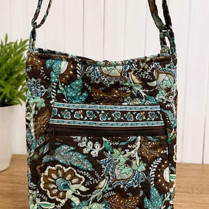 May include: A quilted crossbody bag featuring a dark brown base with a multi-colored floral and paisley pattern in shades of teal, light blue, and cream. The bag includes an adjustable shoulder strap and a horizontal zippered front pocket.