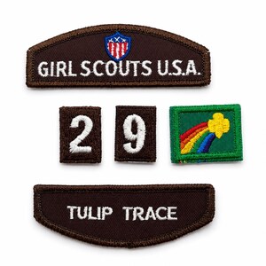 May include: A collection of embroidered patches including a brown Girl Scouts U.S.A. arc patch with a shield emblem, two small brown square patches with the white numbers 2 and 9, a green rectangular patch with a rainbow and yellow flower, and a brown arc patch with the text TULIP TRACE.
