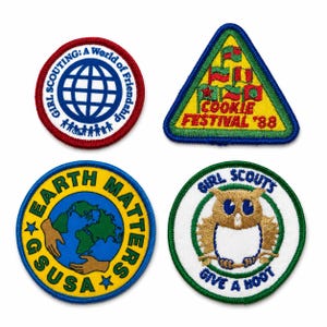 May include: Four embroidered fabric patches. Top left: circular, red border, blue globe, text 'GIRL SCOUTING: A World of Friendship'. Top right: triangular, blue border, yellow background, flags, text 'COOKIE FESTIVAL '88'. Bottom left: circular, blue border, yellow background, globe, hands, text 'EARTH MATTERS GSUSA'. Bottom right: circular, green border, white background, owl, text 'GIRL SCOUTS GIVE A HOOT'.