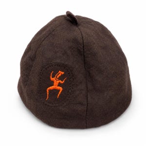 May include: A dark brown felted wool sauna hat with a rounded crown and a small loop at the top. The hat features an embroidered orange graphic of a stylized human figure centered within an oval stitched outline on the front surface.
