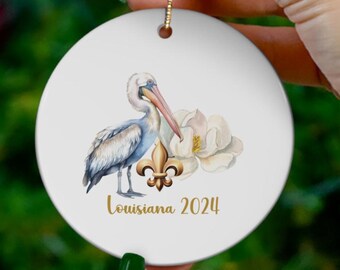 May include: White ceramic Christmas ornament featuring a watercolor illustration of a pelican, magnolia flower, and fleur-de-lis. The text "Louisiana 2024" is printed in gold. The ornament has a gold string for hanging.