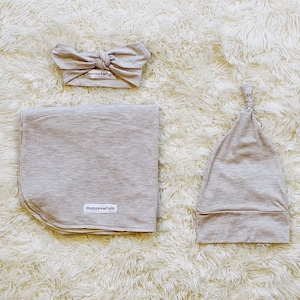 May include: A light beige heathered fabric set consisting of a knotted headband, a rectangular swaddle blanket, and a knotted beanie hat. Each item features a small white rectangular label with the text moose+whale in black lettering.