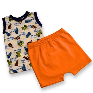May include: A two-piece baby outfit with a sleeveless white vest featuring a colourful truck print and orange shorts.