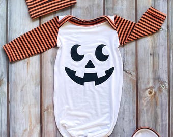 May include: A white baby sleeper and matching hat set with a Halloween pumpkin face design. The sleeper has orange and black striped sleeves and trim. The hat is orange and black striped. The set is made of soft material.