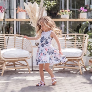 Young girl twirling a sleeveless Summer dress featuring a large vintage inspired palm leaf design with peachy orchids and creamy roses. Think vintage palm springs with a touch of the tropics. Square shaped back and a full twirl skirt