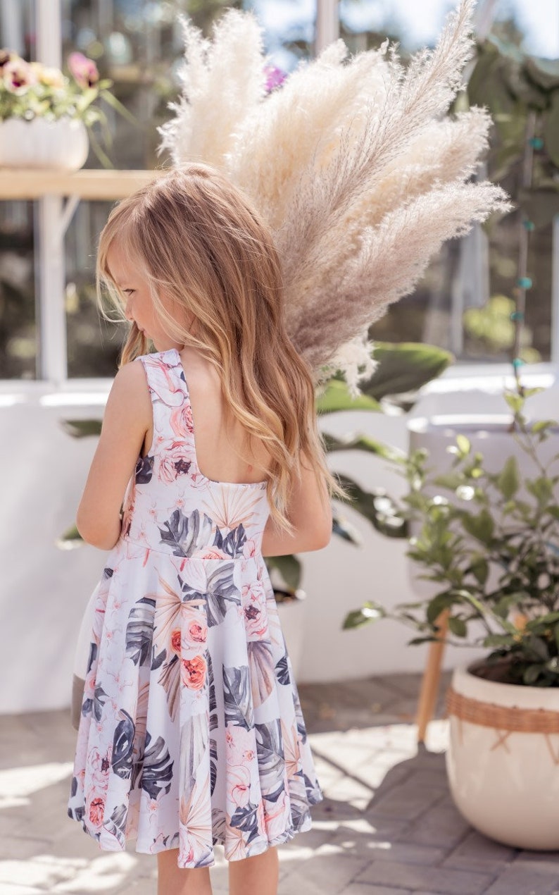 Young girl twirling a sleeveless Summer dress featuring a large vintage inspired palm leaf design with peachy orchids and creamy roses. Think vintage palm springs with a touch of the tropics. Square shaped back and a full twirl skirt