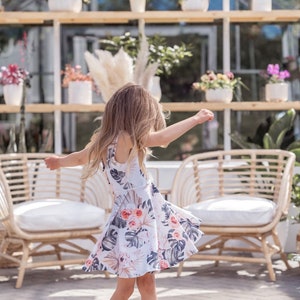 Young girl twirling a sleeveless Summer dress featuring a large vintage inspired palm leaf design with peachy orchids and creamy roses. Think vintage palm springs with a touch of the tropics. Square shaped back and a full twirl skirt