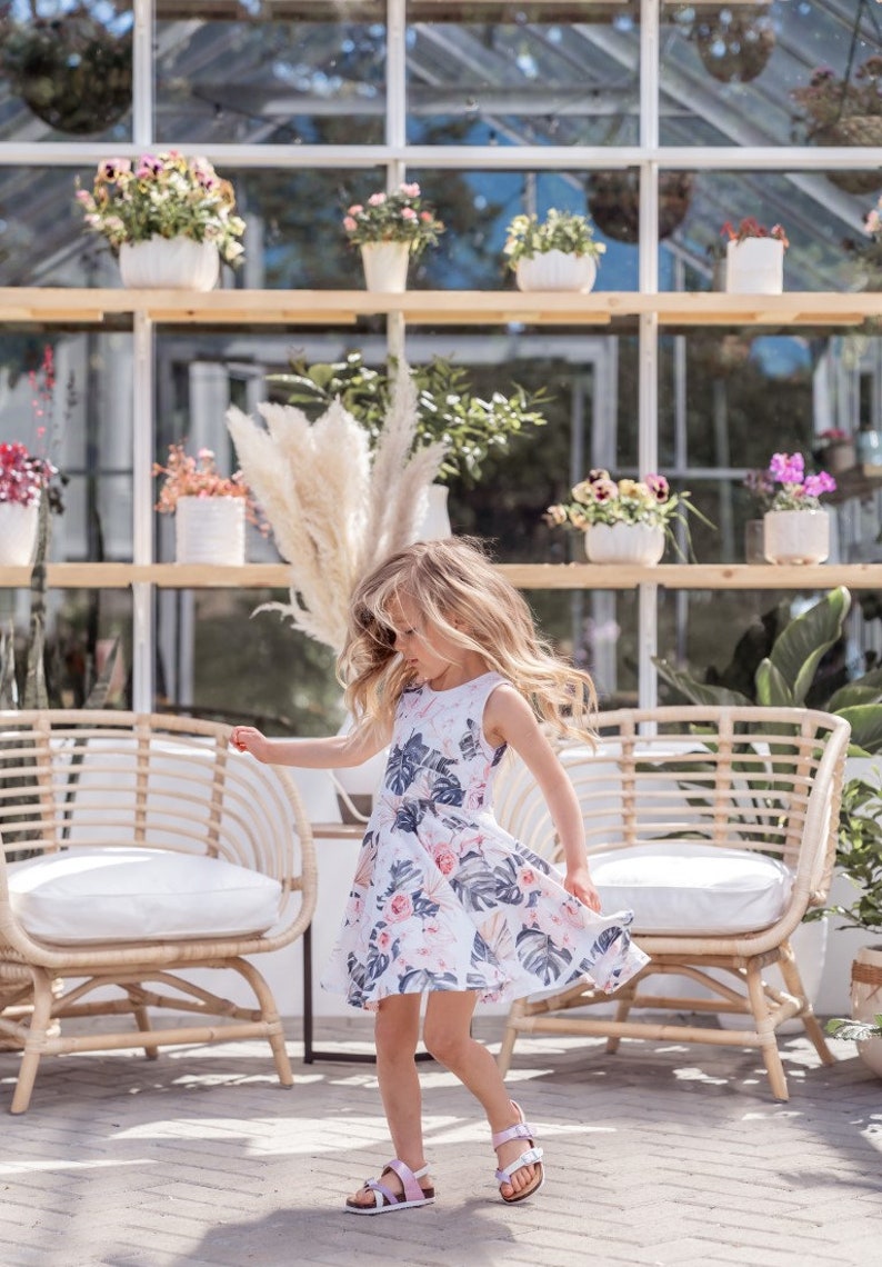 Young girl twirling a sleeveless Summer dress featuring a large vintage inspired palm leaf design with peachy orchids and creamy roses. Think vintage palm springs with a touch of the tropics. Square shaped back and a full twirl skirt