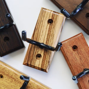 May include: Six wooden guitar wall mounts with black metal hooks. The mounts are in various wood tones, including dark brown, light brown, reddish brown, and a light wood with dark streaks.