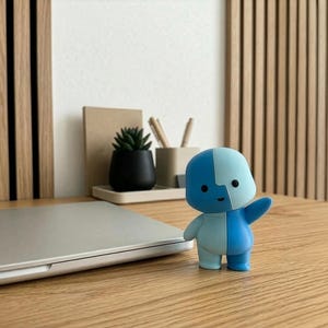 May include: A two-toned blue figurine with a raised arm stands on a wooden desk next to a closed silver laptop. A small black potted plant and pencils are visible in the background. The desk is made of wood.