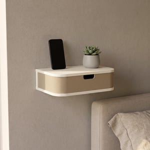 May include: A wall-mounted floating shelf with a white frame and a beige, horizontally ribbed drawer front. A black smartphone stands upright on the top surface next to a small, light grey pot containing a green succulent plant.
