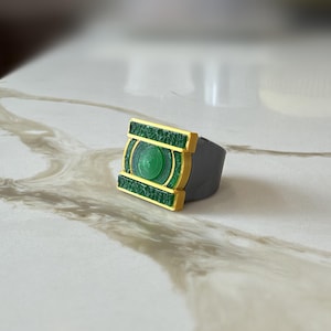 May include: A ring featuring a rectangular face with a central circular green emblem, framed by gold-colored borders and textured green panels. The band of the ring is a solid gray color, providing a contrast to the detailed, multi-colored face.