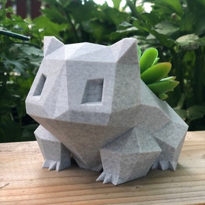 Peut inclure: Un pot de fleurs gris, géométrique, imprimé en 3D en forme de personnage de Pokemon. Le pot contient une petite plante succulente.