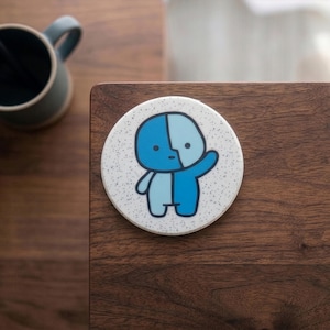 Peut inclure: Un dessous de verre rond et blanc moucheté avec un dessin de personnage de dessin animé. Le personnage est divisé verticalement, avec un côté bleu clair et l'autre bleu foncé. Le personnage fait signe d'un bras.
