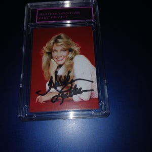 Puede incluir: Fotografía firmada de Heather Locklear en una funda protectora. La foto muestra a Locklear con cabello rubio, un suéter blanco y un fondo rojo. La firma es con tinta negra.