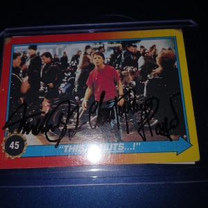 May include: A trading card featuring a scene from a movie, with a man in a red shirt as the central figure. The card has a blue, yellow, and red border, with text and an autograph. The number 45 is visible.