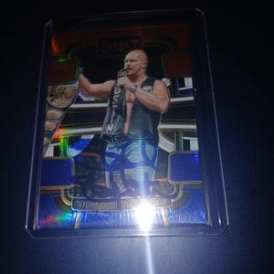 May include: A trading card featuring a wrestler in a black vest and jeans, holding a microphone. The card has a holographic design with the text "STONE COLD STEVE AUSTIN". The card is encased in a protective sleeve.