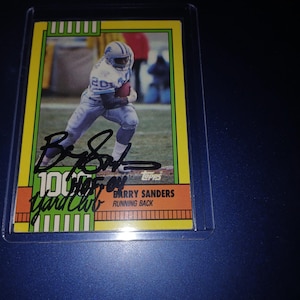 May include: A vintage Topps football card featuring Barry Sanders in a Detroit Lions uniform. The card has a yellow and green border, with Sanders' signature in black ink. Text includes "100 Yard Club" and "Running Back".