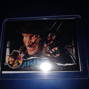 Op de afbeelding: Een verzamelkaart met Freddy Krueger uit "A Nightmare on Elm Street". De afbeelding toont Kruegers littekengezicht, hoed en mes-handschoen. De kaart zit in een beschermhoes.
