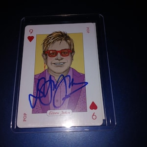 Op de afbeelding: Een speelkaart met een portret van Elton John met een rode bril en een paars pak. De kaart is gesigneerd met blauwe inkt en heeft het nummer 6 en een hartsymbool. Het woord "POP" staat op de kaart.