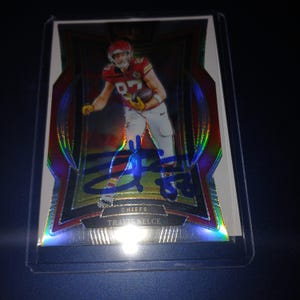 Puede incluir: Una tarjeta de fútbol coleccionable con Travis Kelce de los Kansas City Chiefs. La tarjeta tiene un diseño rojo y plateado con una imagen de Kelce en acción, su firma y el nombre del equipo.