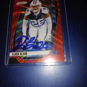 May include: A red and black football trading card with a holographic finish. The card features a photo of Daron Bland, a Dallas Cowboys player, wearing a blue jersey with the number 26. The card has a blue signature and a blue star logo.