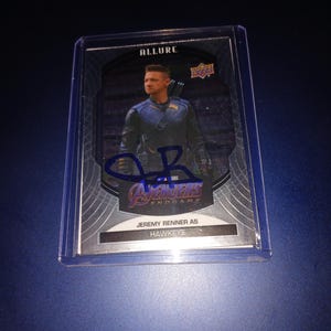 May include: A silver and black trading card featuring Jeremy Renner as Hawkeye from the movie Avengers: Endgame. The card has a silver border and a black background with a silver and black image of Hawkeye. The card also features a blue signature of Jeremy Renner.