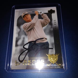 Op de afbeelding: Een golf trading card met een golfer in actie, gesigneerd. De kaart heeft een gouden embleem en de tekst "Legends of the Links". De kaart zit in een beschermende plastic hoes.