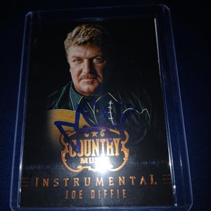 May include: A trading card featuring Joe Diffie, a country music artist. The card has a portrait of Diffie with a guitar, and his signature in blue ink. The card's design includes the words "Country Music" and "Instrumental".